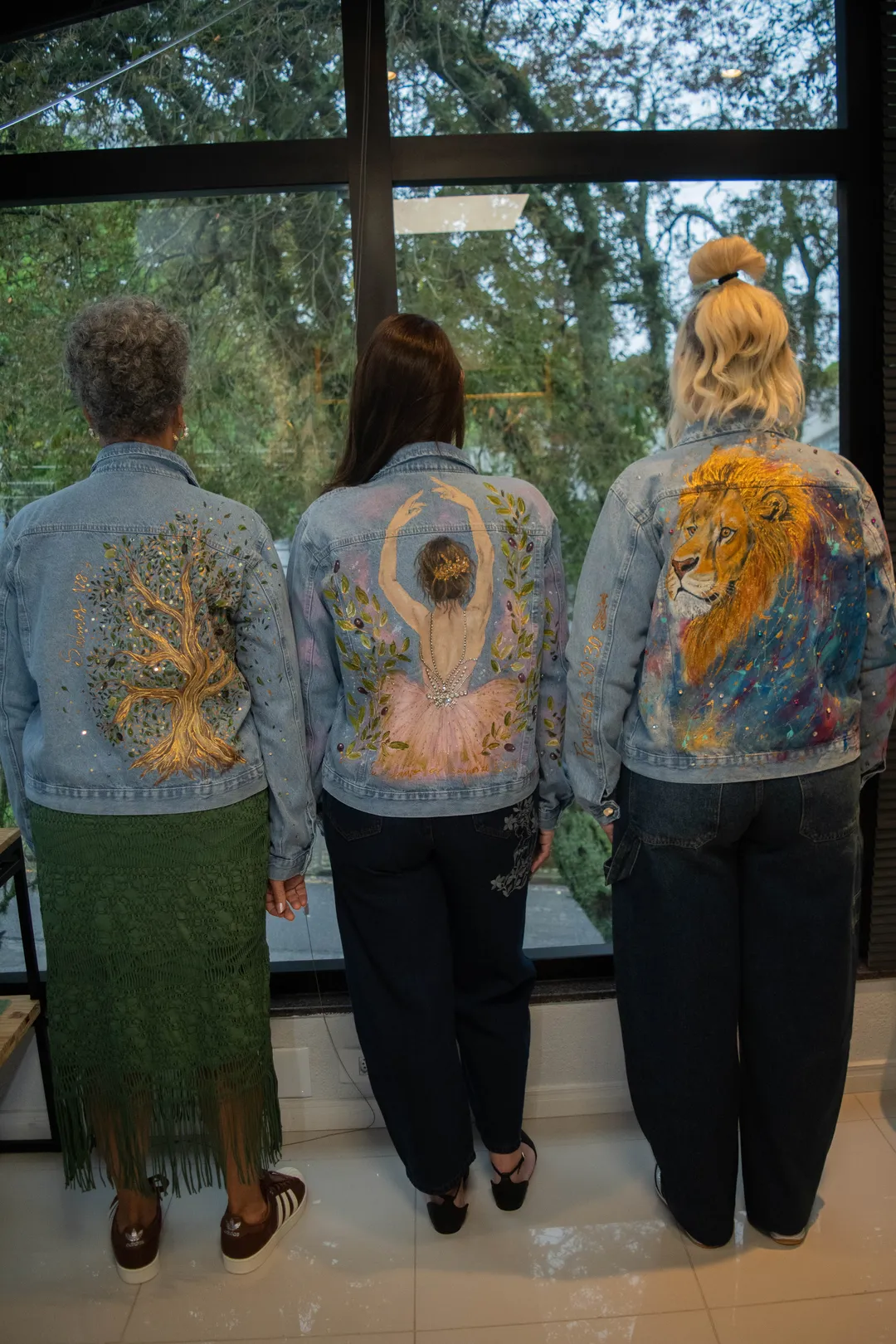 Três mulheres de costas, lado a lado diante de uma janela, vestindo jaquetas R U T H. com pinturas de árvore, bailarina e leão.
