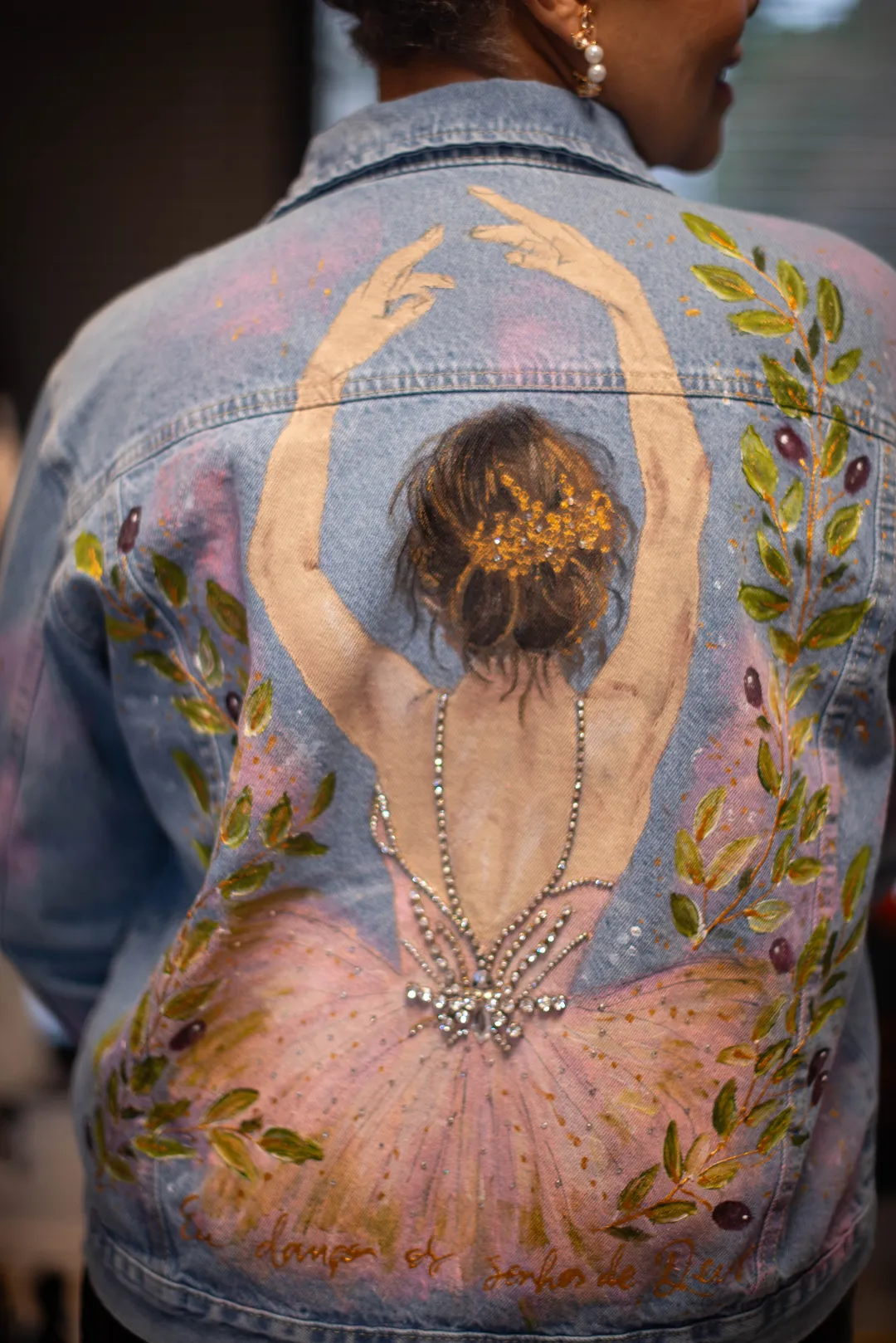 Costas de uma jaqueta jeans com pintura autoral de bailarina de saia rosada, ramos de oliveira e pedrarias.