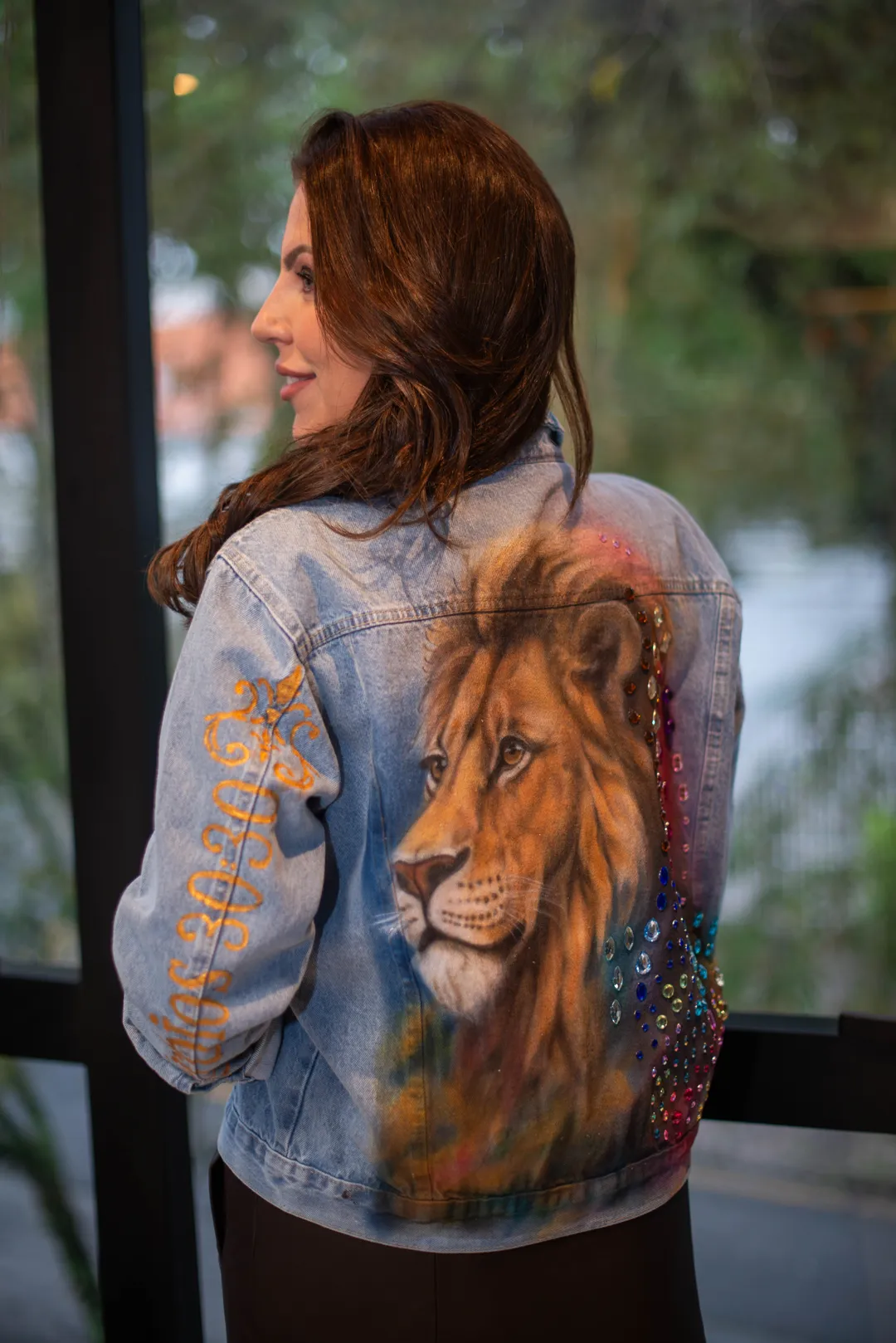 Mulher olhando de lado, vestindo jaqueta jeans com pintura de leão e pedrarias coloridas, com inscrição dourada na manga.