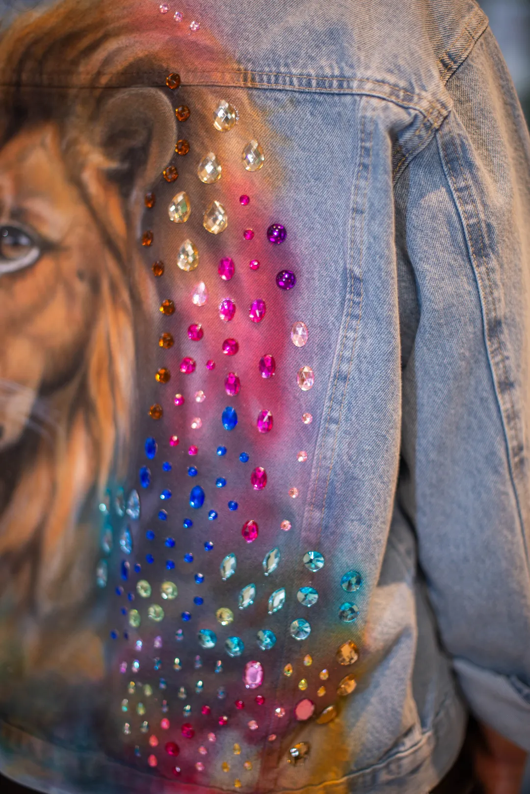 Close de pedrarias coloridas aplicadas ao lado de uma pintura de leão em jaqueta jeans.