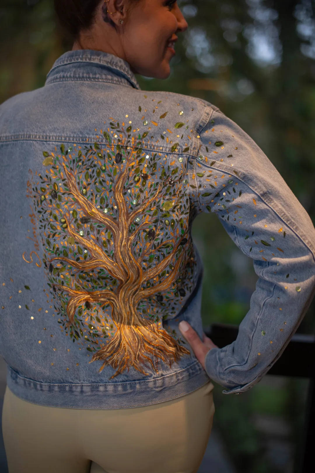 Detalhe da pintura de árvore com tronco dourado e folhagem com pedrarias nas costas de uma jaqueta jeans.