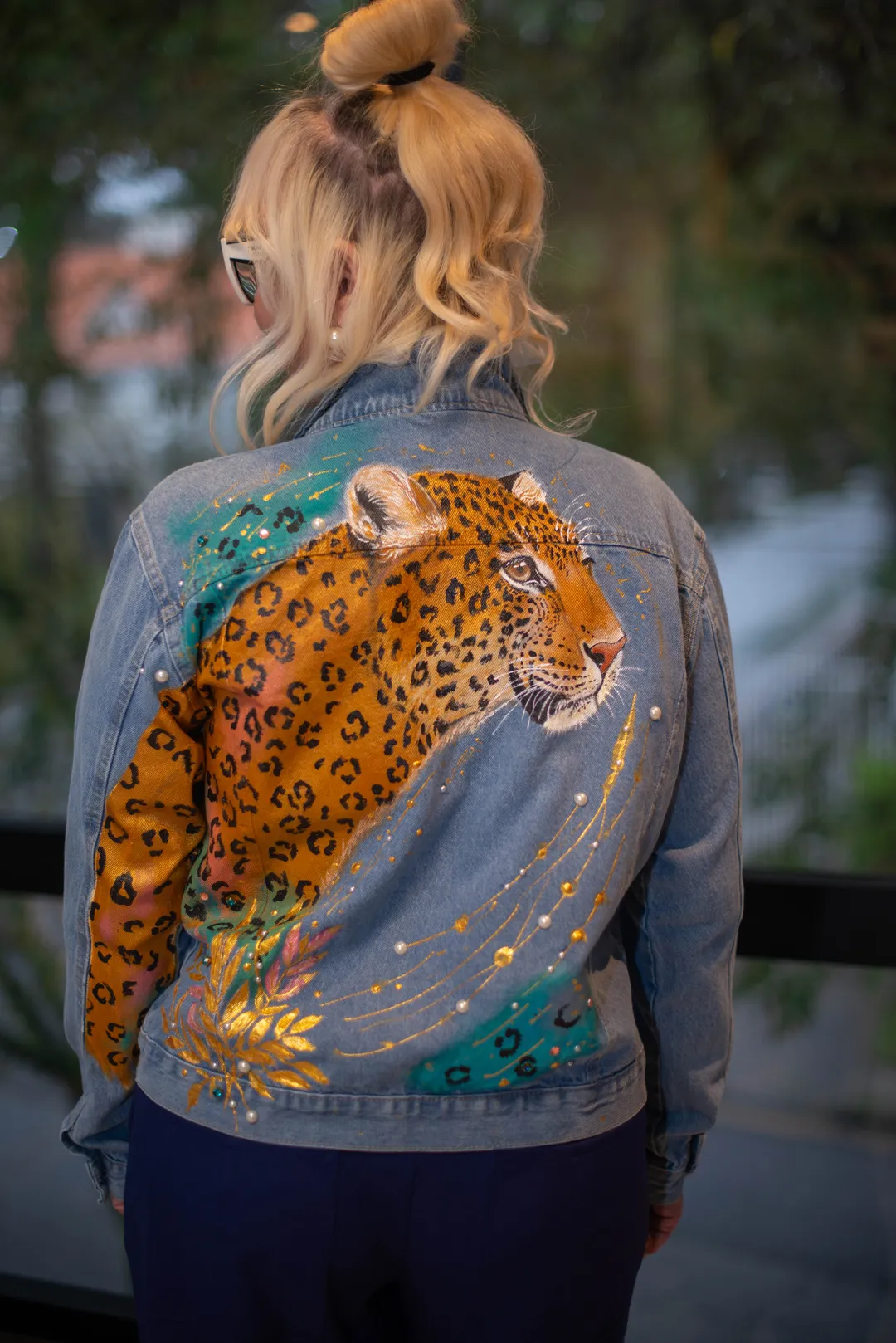 Mulher de costas vestindo jaqueta jeans com pintura de onça em tons dourados, detalhes verde-água e brilhos.