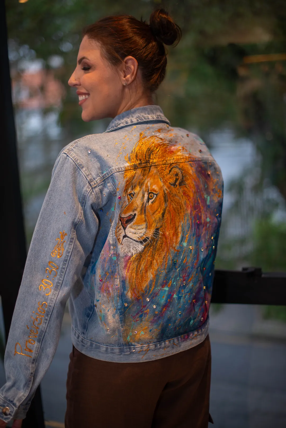 Mulher sorrindo, olhando por cima do ombro, vestindo jaqueta jeans R U T H. com pintura de leão dourado nas costas.