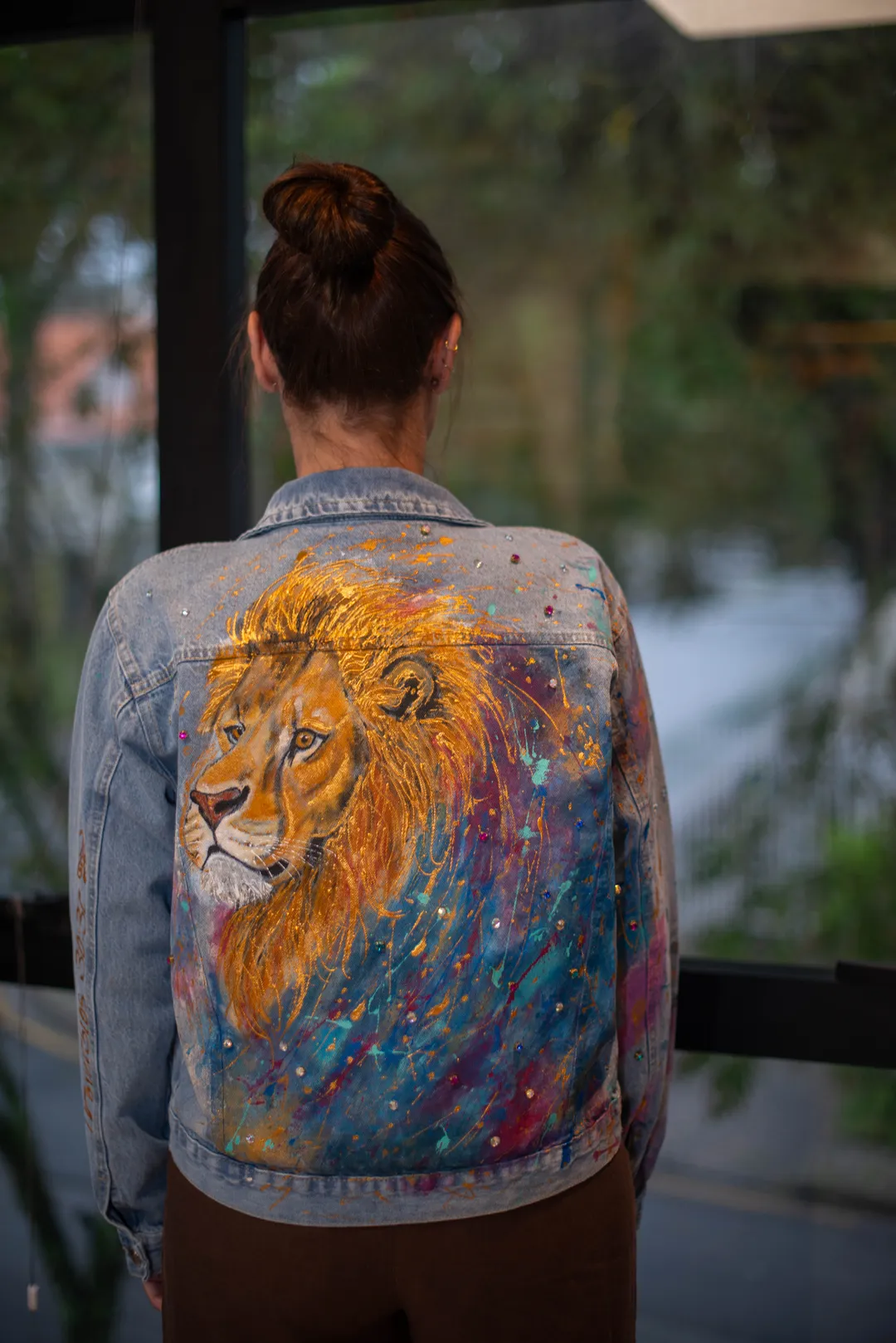 Mulher de costas vestindo jaqueta jeans com pintura de leão em tons dourados sobre fundo de pinceladas azuis, rosadas e lilases.